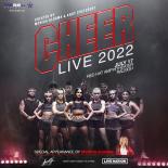 A photo of 13 gymnast posing in black and glitter leotards on stage with a grey fog background. the words cheer live 2022 are above them